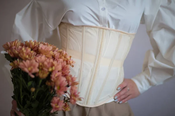 Beyaz gömlekli ve bej korseli kız ellerinde çiçekler tutuyor.