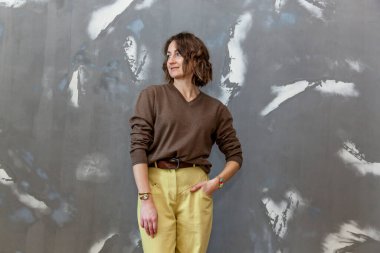 Sarı pantolon ve gri bir duvara karşı kahverengi kazak giyen şık ve şık bir kız.