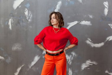 Turuncu pantolonla poz veren şık ve şık bir kız. Gri bir duvara karşı kırmızı bir kazak.