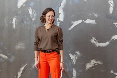 Turuncu pantolonla poz veren şık ve şık bir kız. Gri bir duvara karşı kahverengi bir kazak.