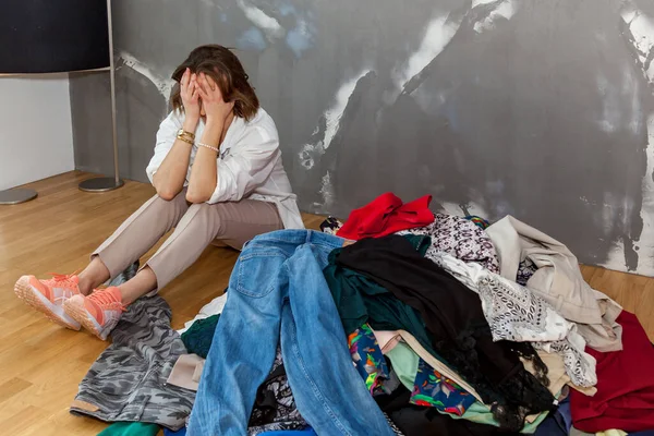 A girl sitting on the floor sorts out her wardrobe, a mountain of things in the room against a gray wall. The girl sorts out her wardrobe
