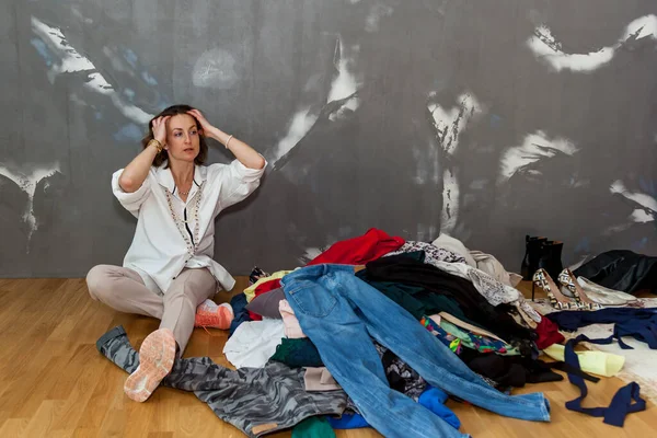 A girl sitting on the floor sorts out her wardrobe, a mountain of things in the room against a gray wall. The girl sorts out her wardrobe