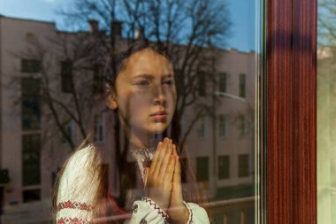 A girl in an embroidered dress prays near the window. The girl protests against the war, and shows patriotism and love for Ukraine. Ukrainian-Russian conflict, war in Ukraine