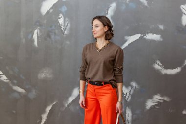 Turuncu pantolonla poz veren şık ve şık bir kız. Gri bir duvara karşı kahverengi bir kazak.