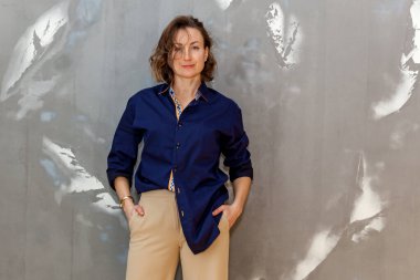 Gri bir duvara karşı bej pantolon ve mavi gömlekle poz veren şık ve şık bir kız.