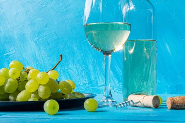 Doğada bir şişenin yanında, yeşil üzümlü, ahşap, mavi bir masada beyaz şarap..