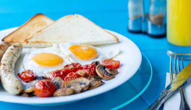 Kızarmış yumurta, ızgara sosis, kızarmış mantar, vişneli domates ve ahşap mavi masada beyaz bir tabakta bir bardak portakal suyu ile çıtır tost..