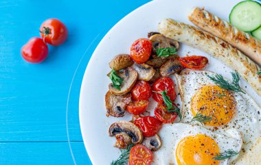 Kızarmış yumurta, ızgara sosis, kızarmış mantar ve vişneli domates, ahşap bir masada beyaz tabakta taze salatalık dilimleri. Yukarıdan görüntüle.