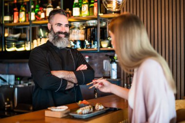 Genç kadın suşi restoranında suşi yiyor ve yemek çubuklarıyla suşi tadıyor. Suşi ustası memnun çünkü yemeği seviyor..