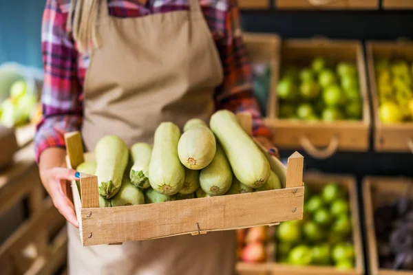 Woman works in fruits and vegetables shop. She is holding basket with marrow squash.