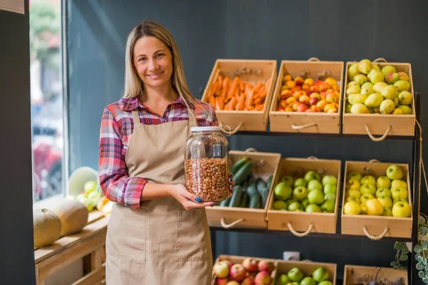 Woman works in fruits and vegetables shop. She is holding jar with almond.