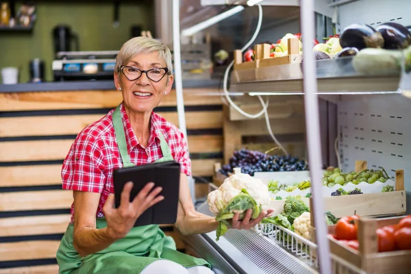 Mature woman works in fruits and vegetables shop. She is examining goods.