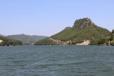 Sakin yeşil-mavi deniz açık bir yaz gününde, alçak tepelerle çevrili bitki örtüsü, bulutsuz mavi gökyüzü