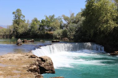 Parlak turkuaz bir göl üzerinde alçak köpüklü bir şelale etrafını kayalar ve ağaçlarla çevrili berrak bir yaz gökyüzü altında