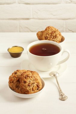 Küçük tabaklar üzerinde ceviz ve kuru üzümlü yulaf ezmesi kurabiyeleri ve beyaz tuğla bir duvara karşı beyaz ahşap bir masanın üzerinde duran bir fincan siyah çay. Yakın çekim..