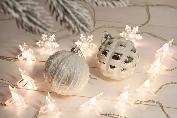 白色和银色的圣诞球躺在一个白色的木制桌子上 旁边是一个明亮的花环 透过松树枝条观看 — 图库照片