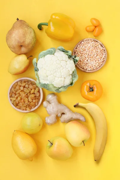 Yellow fruits and vegetables on a yellow background. Pears, raisins, lemon, bergamot, apples, ginger root, paprika, cauliflower, kumquat, peeled dried peas, tomato, banana. Top down view. Closeup.