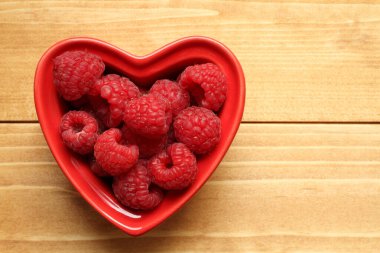 Raspberries lie in a heart-shaped ceramic bowl, which stands on a wooden table. View from above