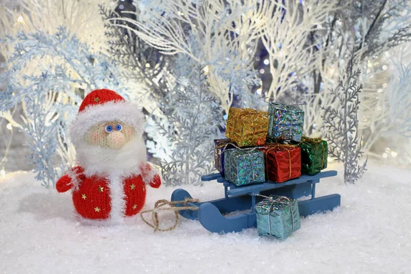 A knitted toy Santa stands on the artificial snow and a toy sled with gifts stands next to him against the backdrop of white and blue artificial trees. Closeup. Night studio shooting.