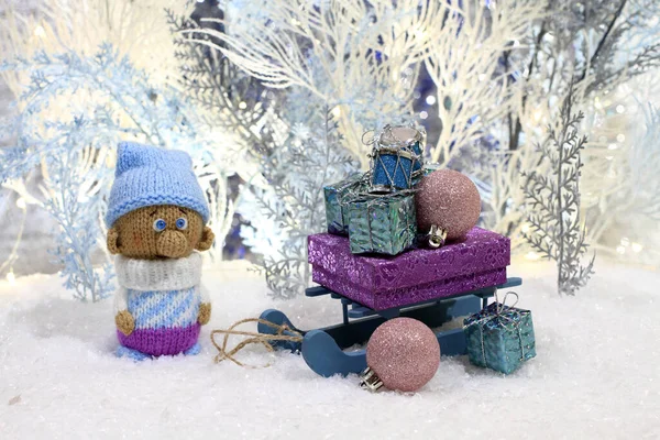 A knitted toy gnome stands on the artificial snow and a toy sled with gifts stands next to him against the backdrop of white and blue artificial trees. Closeup. Night studio shooting,
