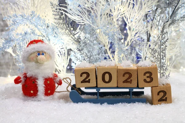 A knitted toy Santa stands on the artificial snow and a toy sledge with wooden cubes with the numbers 2023 stands next to him against the backdrop of blue and white artificial trees. Closeup. Night studio shooting.