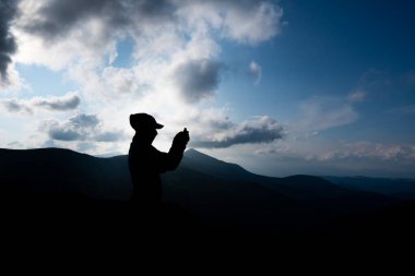 Gün batımında dağların tepesinde cep telefonu tutan insan elleri silueti. Erkek telefonunda fotoğraf çekiyor. Güneşin parıltısı silueti aydınlatır..