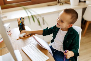 Öğrenci çocuk odasında, masasında ders çalışıyor. Çocuk dizüstü bilgisayar kullanıyor ve deftere yazı yazıyor. Kitaplar ve tablet masanın üstünde. Qurantine sırasında evde çalış. Yeşil Ekran