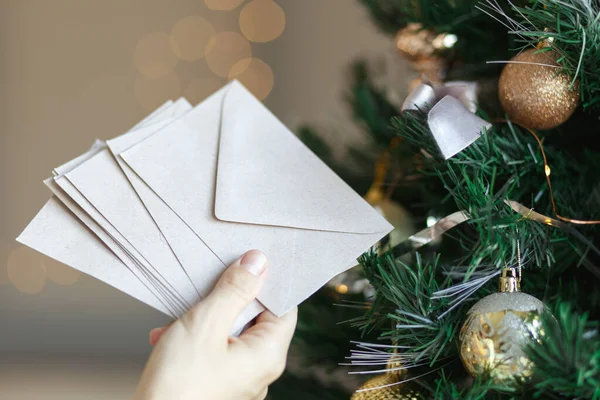 Main tenant pile d'enveloppes en papier artisanal pour la conception d'invitation de carte de vœux de Noël sur fond d'arbre de Noël avec bokeh — Photo