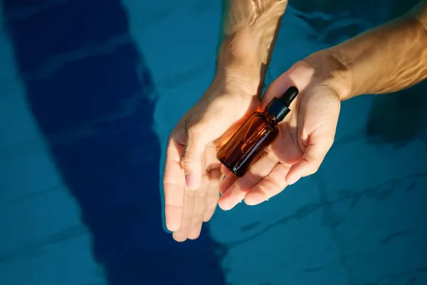 Frau Hand hält leere Bio-Serum-Hautpflegeprodukt Glas Bernstein Flasche für den Einsatz in einem Tag mit einfachen hellblauen Schwimmbad Wasser Hintergrund — Stockfoto