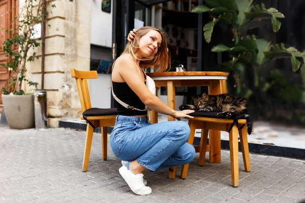 Eine junge Hipsterfrau sitzt an einem Tisch in einer Café-Bar im Freien und spielt mit der Katze. Sommerzeit — Stockfoto