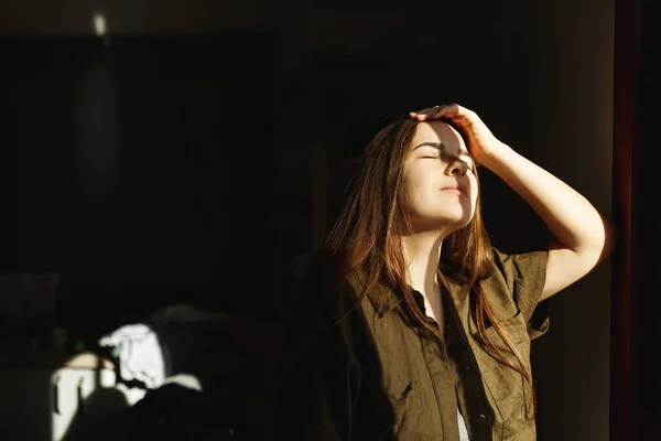 Jolie jeune femme dans l'appartement ouvrant les rideaux de fenêtre après le réveil, profitant du soleil matinal.Concept de santé mentale. Lumière et ombres — Photo