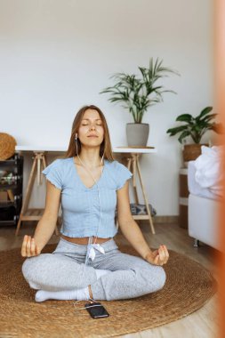 Kulaklık takan, internetten meditasyon yapan, akıllı telefon çalgıcısı uygulamasında müzik dinleyen, evde otururken meditasyon uygulayan, farkındalık sabahı rutininin bir parçası olan genç bir kadın. Huzurlu bir ruh hali.
