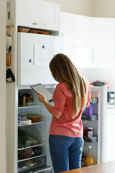 Young woman standing next to an opened refrigerator door, holding a smartphone and ordering fresh fruit and vegetables online for home delivery — Stock Photo, Image