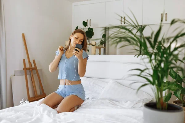 Young teen girl in pajama standing on her bed and check smartphone. Cheerful caucasian adolescent girl with headphones in her bedroom — Stockfoto