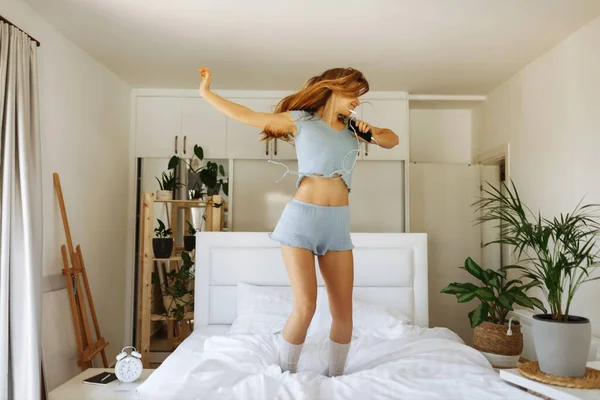 Young teen girl in pajama standing on her bed and singing a song. Cheerful caucasian adolescent girl with headphones dancing on her bedroom — Stockfoto