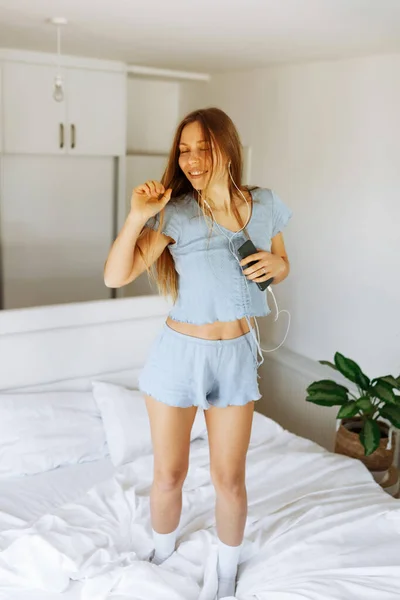 Young teen girl in pajama standing on her bed and singing a song. Cheerful caucasian adolescent girl with headphones dancing on her bedroom — Foto Stock