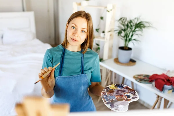 Woman artist holding paintbrush.Creative woman enjoying painting.Young beautiful woman in art studio, taking colorful paints from palette,creating masterpiece on easel, work, imagination. — Fotografia de Stock