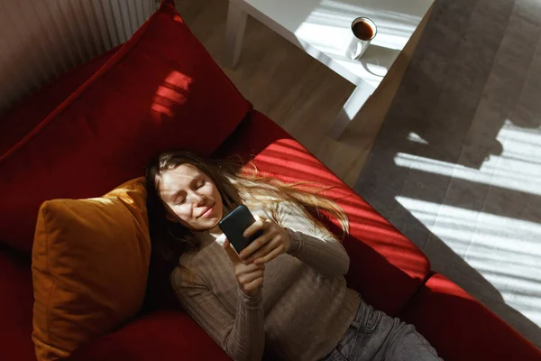 Young woman using smartphone at cozy home on sofa in living room, top view, sunlight from window