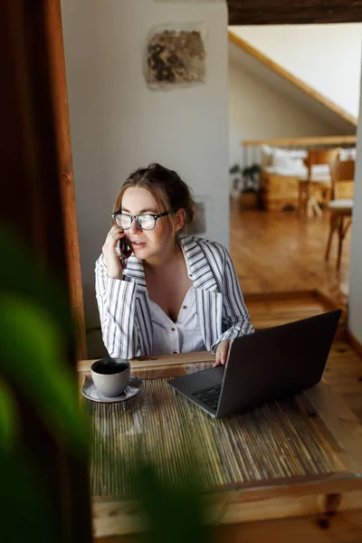 Happy young businesswoman sitting at table with laptop, holding mobile phone call with client in office. Skilled manager saleswoman calling customer, giving professional consultation.
