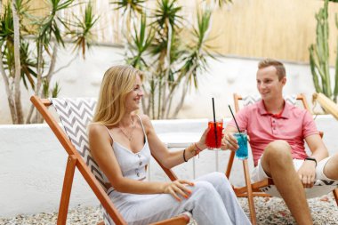 Happy young couple with glasses of lemonade sitting on deck chairs at sea beach or cafe place. 