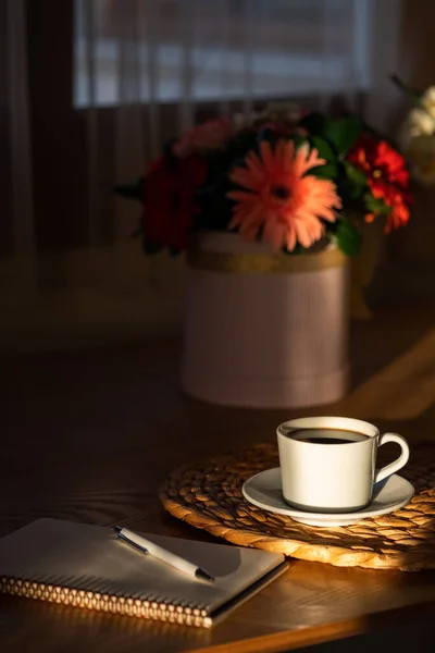 Cozy breakfast with cup of coffee, flowers and open notebook on rustic wooden table. Morning sunshine with shadows