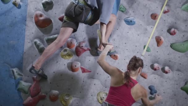 Sport flicka och atletisk man klättra klätterväggen i gymmet. Mannen ger en hjälpande hand, hjälper till att resa sig. Använder ett säkerhetslina på boulderingväggen — Stockvideo