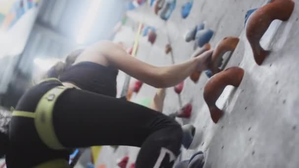 Sportowa dziewczyna wspina się na ścianę wspinaczkową. Używa liny bezpieczeństwa i ubrań wspinaczkowych. Chwyta się za kamienie i grzbiety — Wideo stockowe