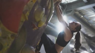 Sporcu kız spor salonundaki tırmanma duvarına tırmanıyor. Kaya duvarında güvenlik ipi kullanıyor. Taşları eliyle tutuyor.