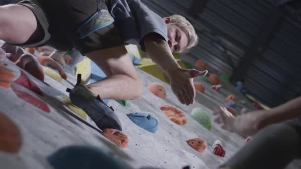 Sport girl and athletic man climb the climbing wall in the gym. The man gives a helping hand, helps to rise. Uses a safety rope on the bouldering wall — Stock Video