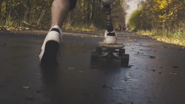 在秋天的森林里，一个身穿金属仿生假肢的年轻人正骑着滑板。滑板上的一条人造腿推开了柏油 — 图库视频影像