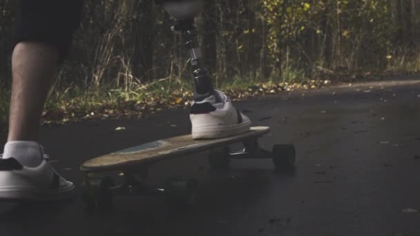 Молодой человек с металлической протезной ногой катается на скейтборде в осеннем парке. Заниматься спортом с искусственной ногой — стоковое видео