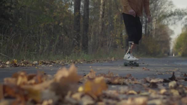 Egy fém műlábú fiatalember gördeszkázik egy őszi parkban. Mesterséges lábú sportolók. — Stock videók