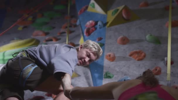 Sport dziewczyna i sportowiec człowiek wspiąć się na ścianę wspinaczkową w siłowni. Podaje pomocną dłoń, pomaga wstać. Używa liny bezpieczeństwa na ścianie głazu. — Wideo stockowe