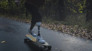 Metal protez bacaklı genç bir adam sonbahar parkında kaykaya biniyor. Yapay bacakla spor yapmaya git.